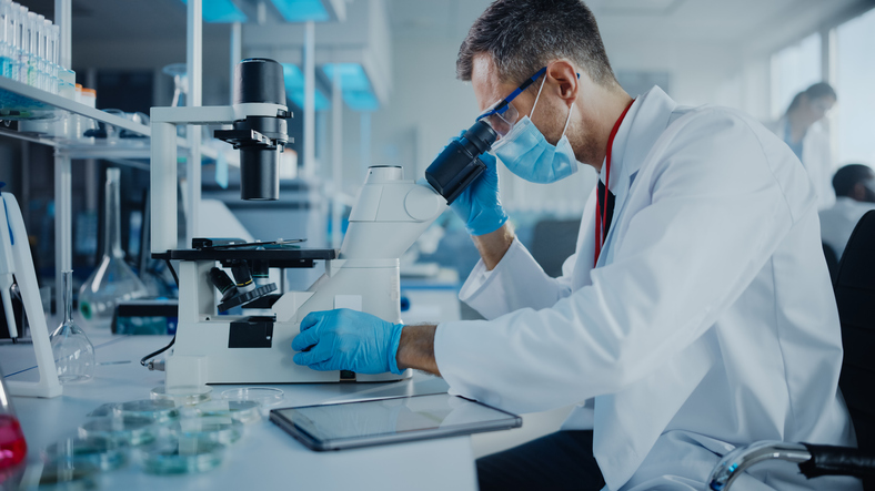 Medical Development Laboratory: Scientist Wearing Face Mask Looking Under Microscope and Using Digital Tablet. Specialists Working on Medicine, Biotechnology Research in Advanced Pharma Lab